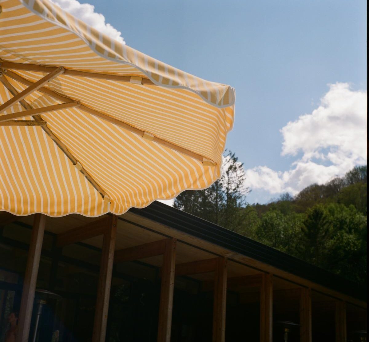 Eastwind Oliverea Valley A striped parasol cuts across the Catskills sky beside Eastwind's low timber pavilion—the kind of cheerful summer detail that immediately tells you this is a mountain retreat, not a wilderness endurance test